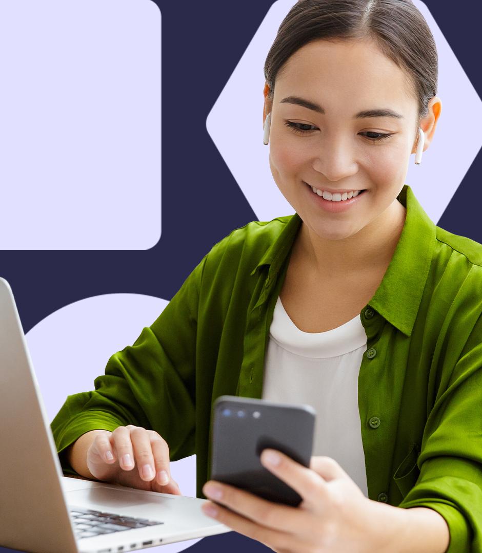 A smiling woman uses a smartphone while working on a laptop, wearing wireless earbuds.