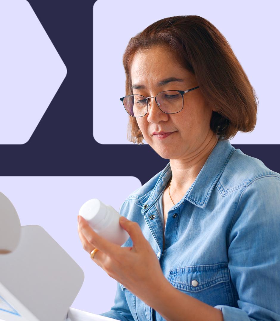 A woman wearing glasses and a denim shirt opening a package and inspecting a white pill bottle, reading the label with focus.
