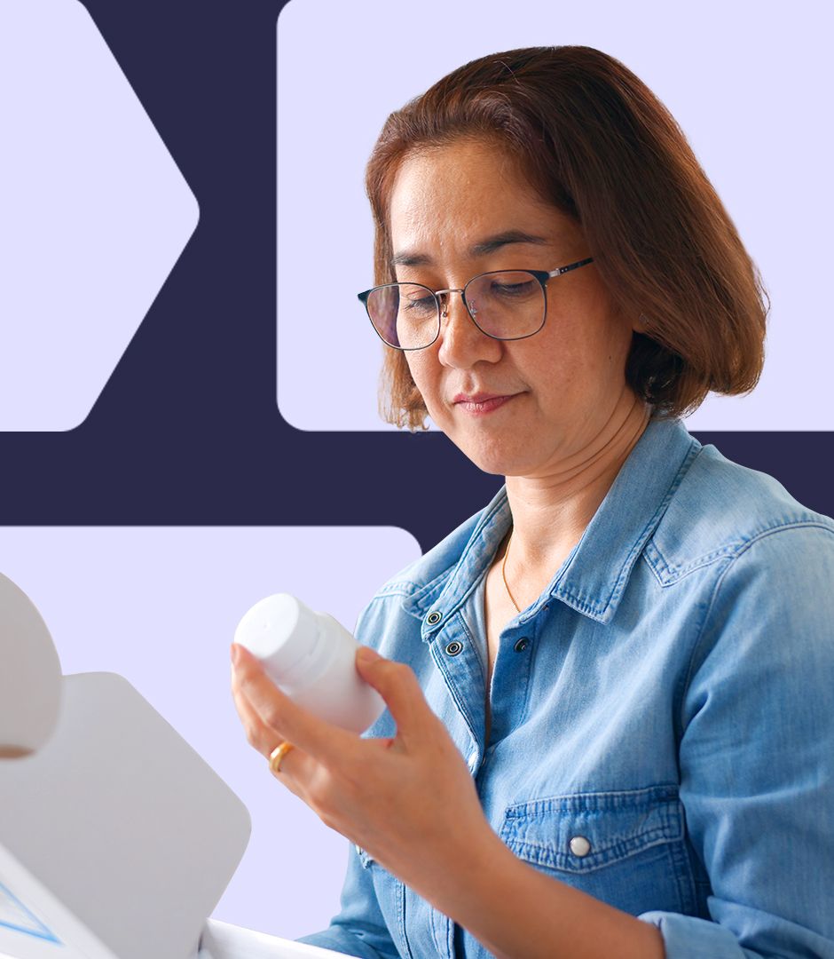 A woman wearing glasses and a denim shirt opening a package and inspecting a white pill bottle, reading the label with focus.