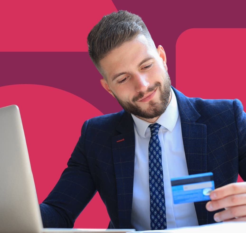 A man wearing a business suit is sitting with an open laptop and looking down at a credit card in his hand.