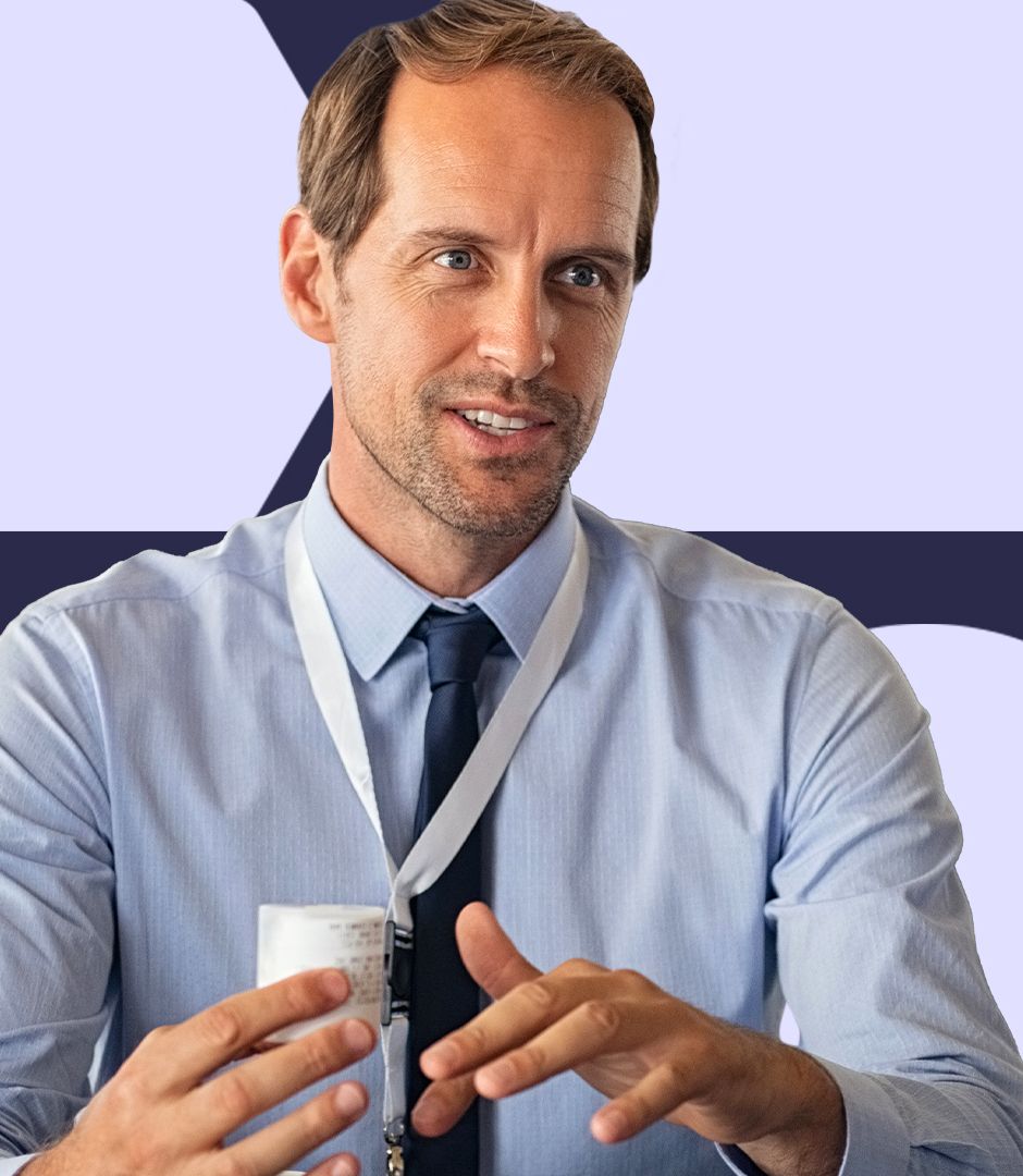 A man in a light blue dress shirt and dark tie, wearing a lanyard, gestures while speaking in a professional setting, holding a small prescription label paper.