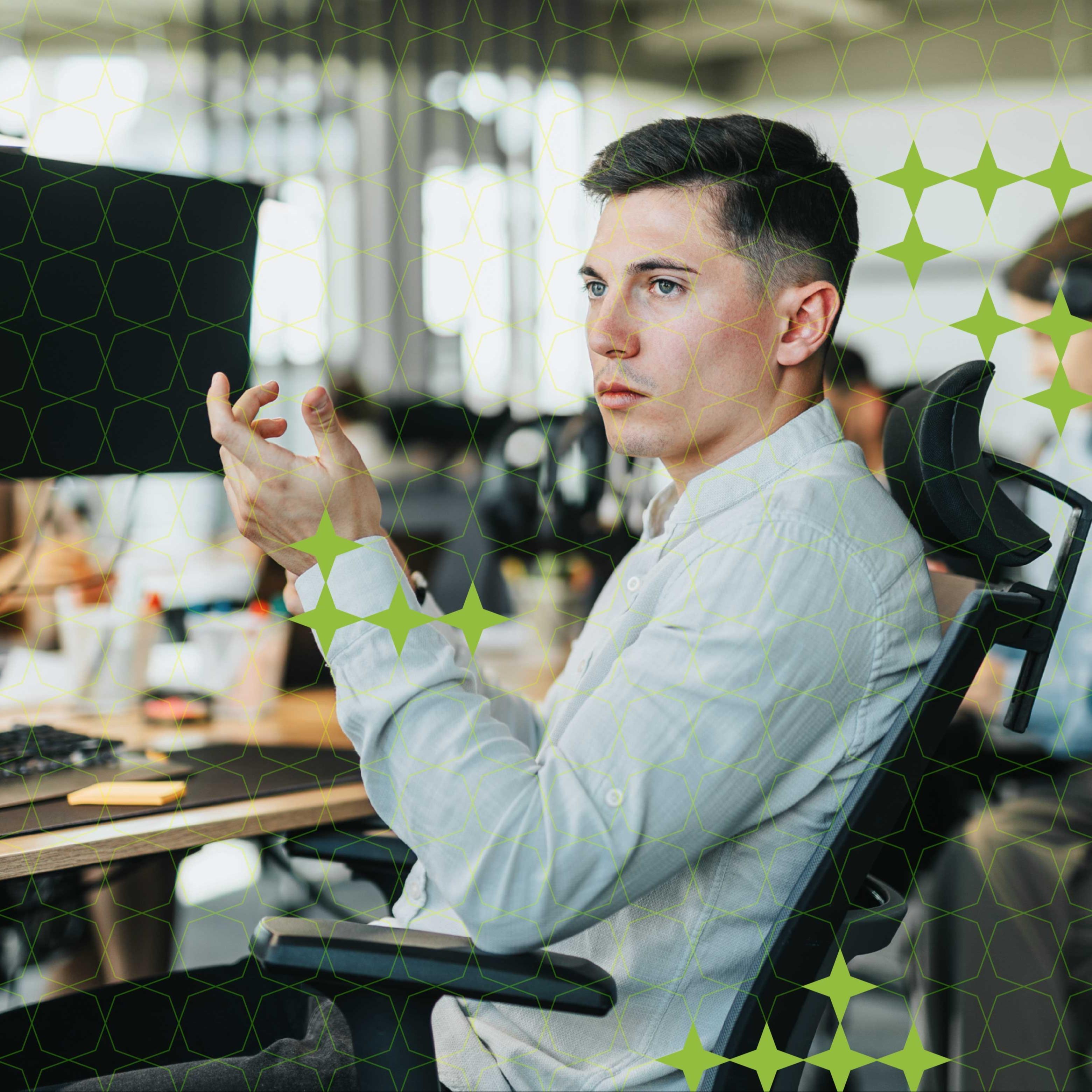 A man sits at a desk with with two monitors in an office, gesturing to a colleague not seen.
