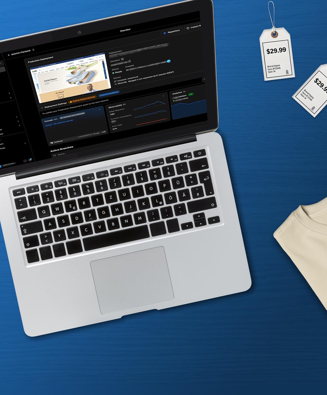Laptop que muestra un panel de control de implementación de producción de e-commerce de Belk, junto a etiquetas de precios minoristas y una camiseta crema doblada sobre un fondo azul.