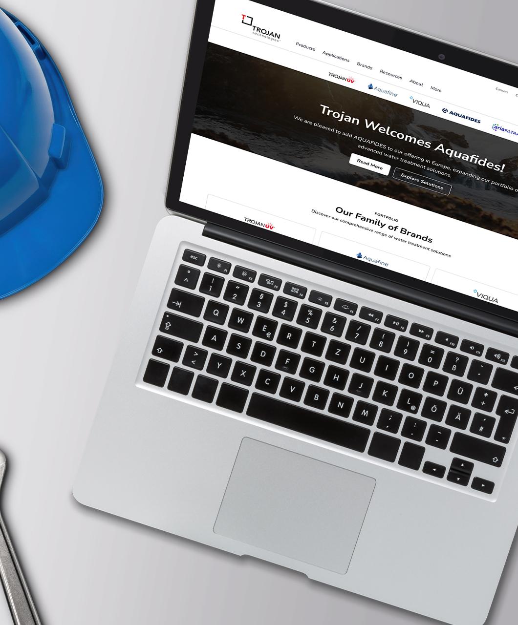 Pantalla de laptop que muestra la página de inicio del sitio web de Trojan Technologies, con un casco de seguridad azul apoyado a su lado sobre una superficie gris claro.