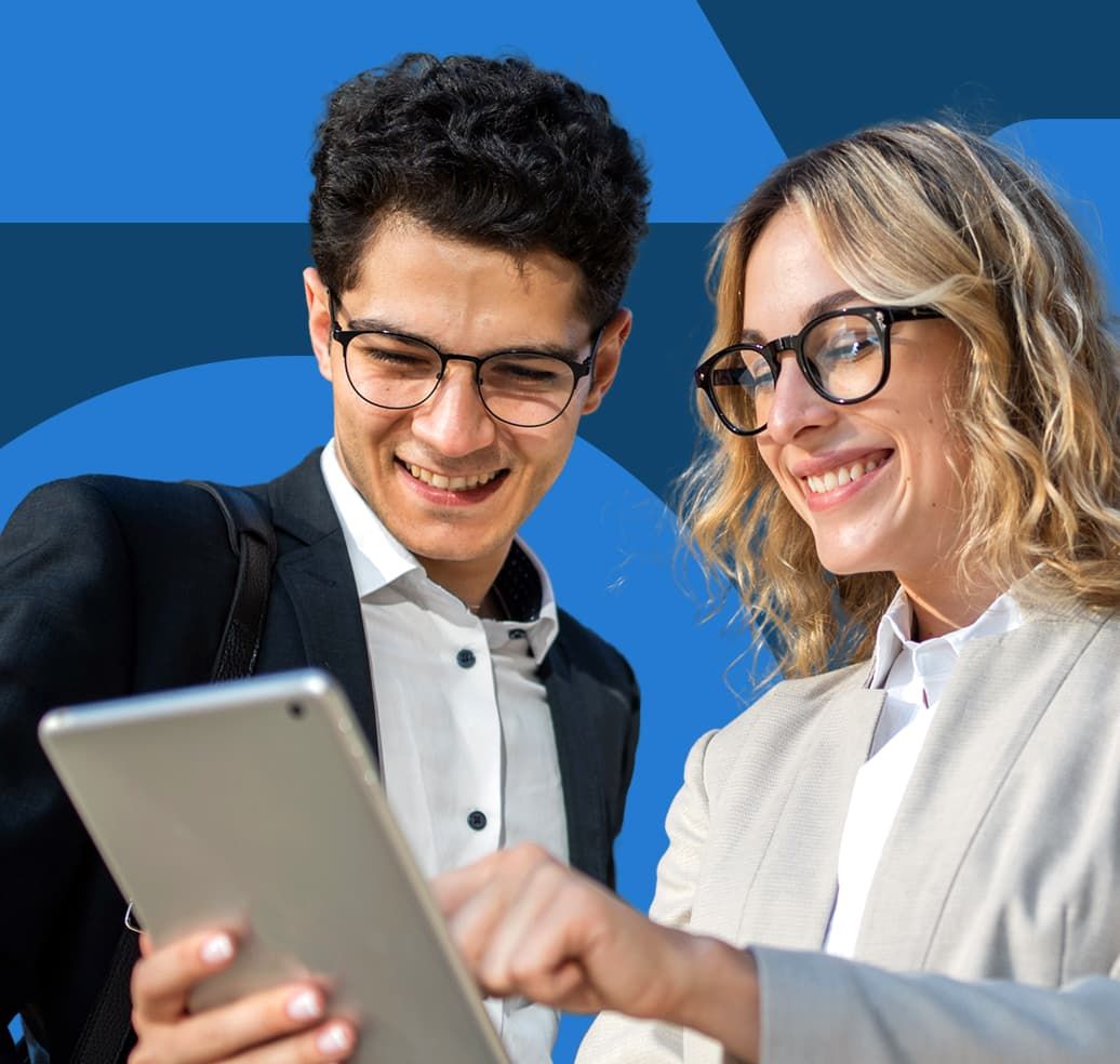 Two colleagues dressed in business attire are standing next to each other and smiling at an iPad screen. The woman holding the iPad is pointing to something on the screen.