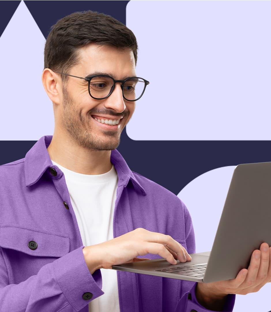 A man wearing a white t shirt and purple button-down shirt is holding a laptop and smiling at the screen.
