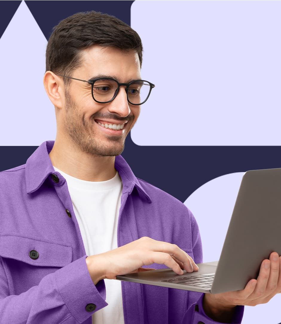A man wearing a white t shirt and purple button-down shirt is holding a laptop and smiling at the screen.