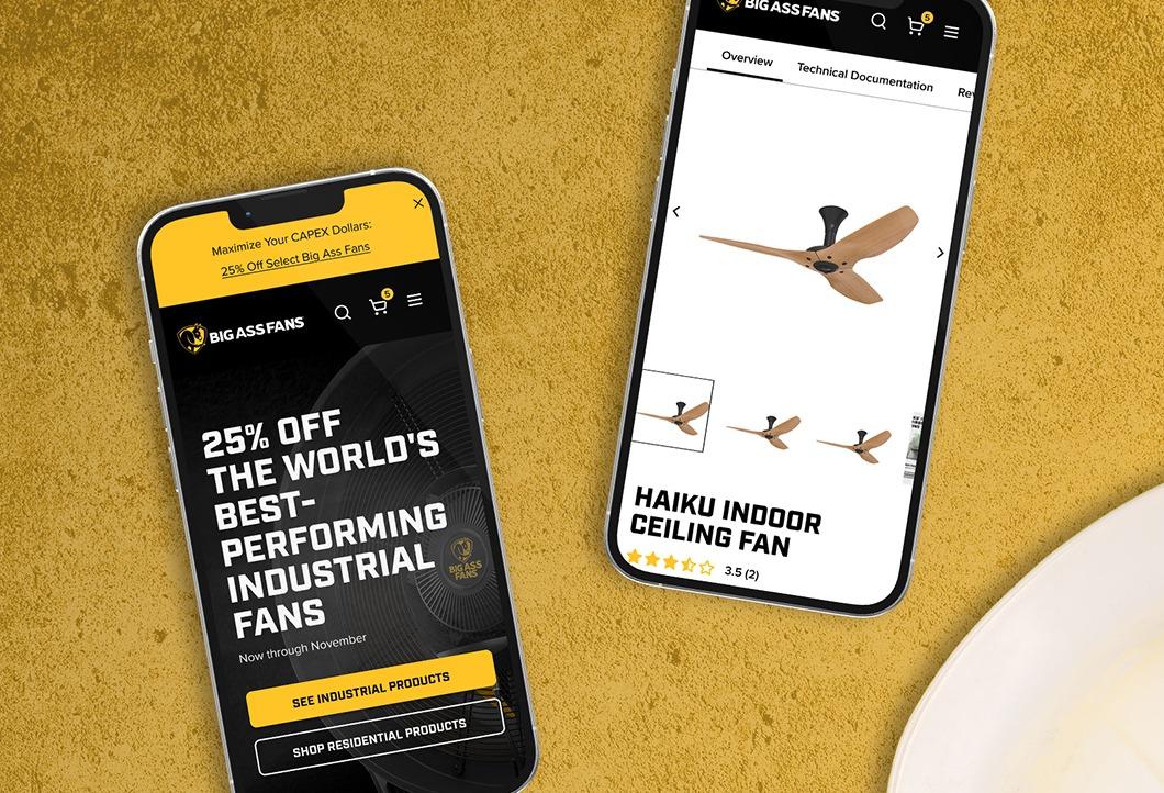 Two phones on a mustard surface show the Big Ass Fans mobile site — a homepage promo and the Haiku Indoor Ceiling Fan product page — beside a branded sticker and a white hard hat.