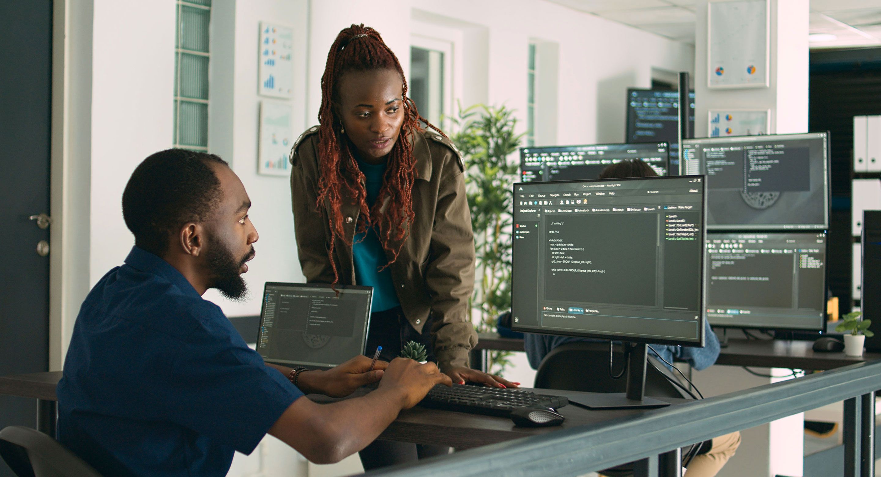 Two developers discuss code while working together at a multi-monitor workstation in a modern office.