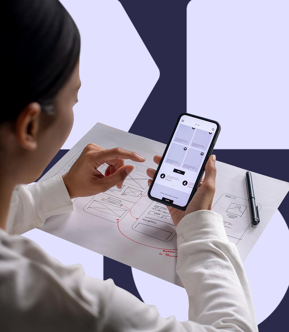 A person views a mobile app prototype on a smartphone over a printed wireframe sketch.
