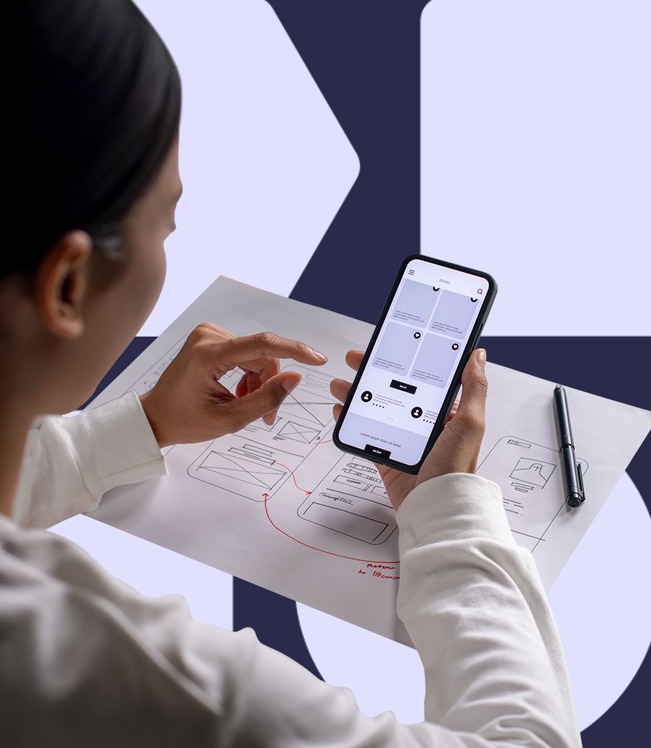 A person views a mobile app prototype on a smartphone over a printed wireframe sketch.