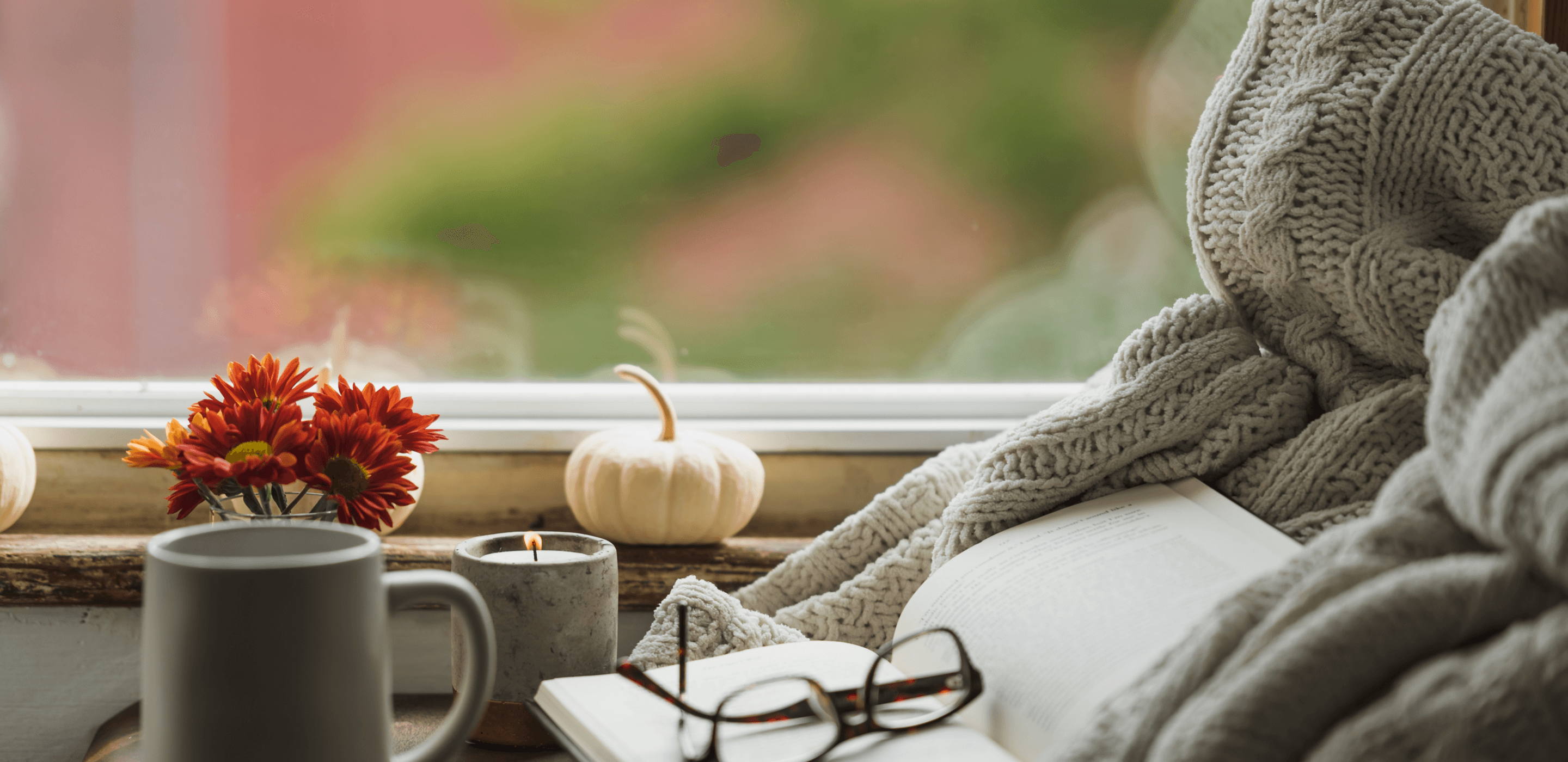 A white pumpkin sitting on a window sill, near a lit candle, a pair of glasses, a book, and a blanket.