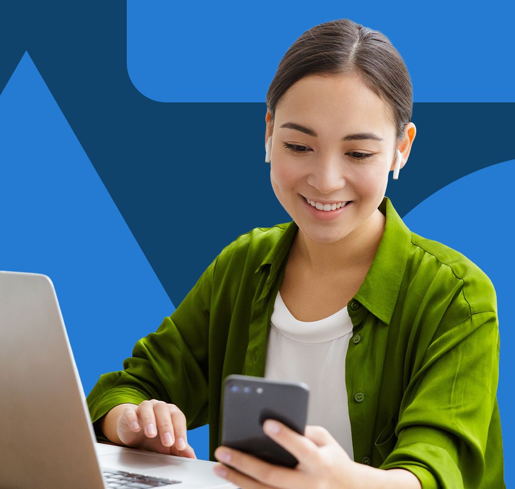 A cheerful woman in a green shirt uses her smartphone while working on a laptop.